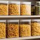 Clear airtight glass storage containers filled with various gluten-free pasta shapes including penne and rotini arranged on organized pantry shelf