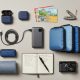 An organized travel flat lay showing creative travel gifts including a power bank, packing cubes, noise-canceling earbuds, and a passport holder on a neutral background